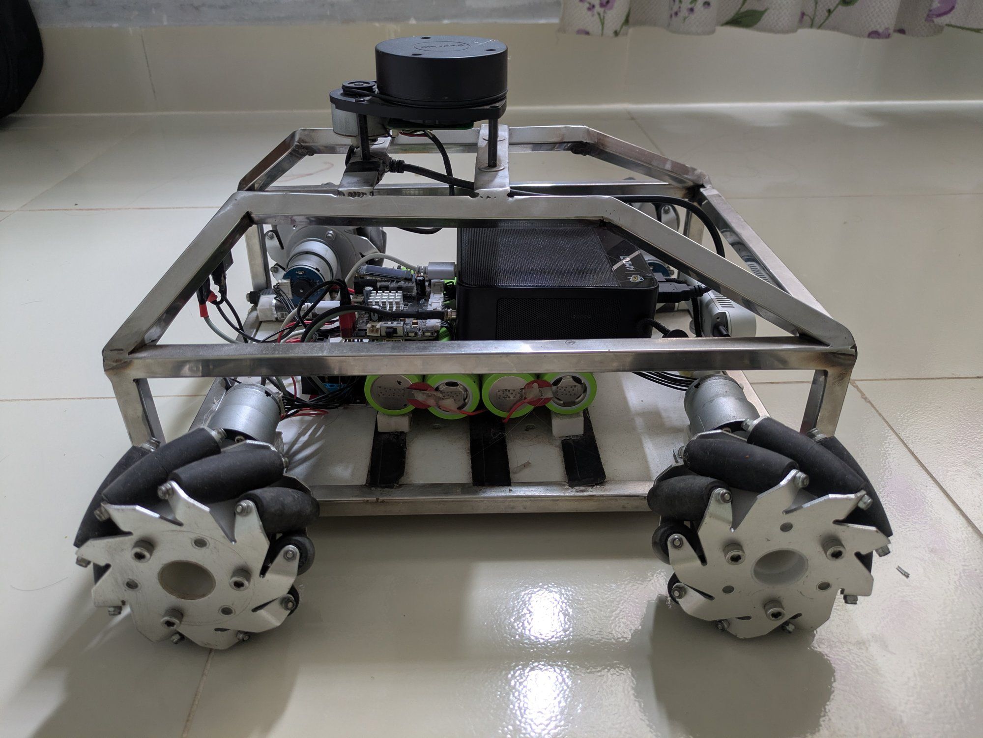 OrbiBot V1 from the side: a welded stainless-steel frame on four mecanum wheels, a LiDAR on top, a mini PC, a motor board and a battery pack inside.